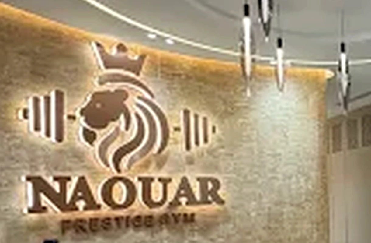 The Naouar Prestige Gym mark — a crowned lion between weight-plate motifs — lit in bronze on a stone wall.